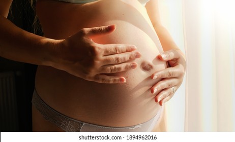 Closeup of pregnant woman standing in sun light and gently stroking her big belly. Concept of happy pregnancy and baby anticipation.