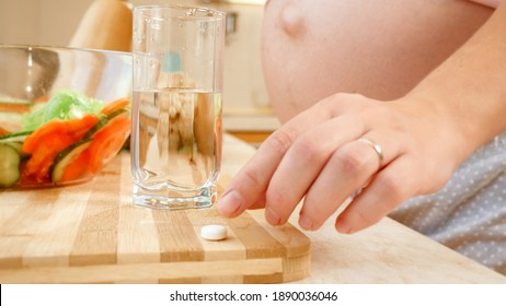 Closeup Of Pregnant Woman Taking Vitamin Pill And Drinking Glass Of Clear Water Before Breakfast On Kitchen. Concept Of Healthy Lifestyle, Nutrition And Hydration During Pregnancy.