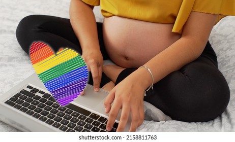 A closeup of a pregnant woman's belly as she works on her laptop with a rainbow 2D heart on top of it