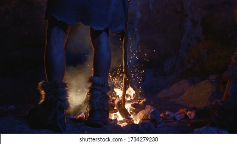 Close-up Prehistoric Tribe Leader Legs Standing At Bonfire Warming Up Near Cave Dwelling. First Human Society. Era Of Ice Age.