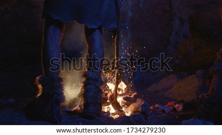 Close-up prehistoric tribe leader legs standing at bonfire warming up near cave dwelling. First human society. Era of ice age.