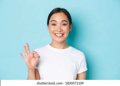 Close-up of pretty satisfied smiling asian girl in white t-shirt, showing recommendation gesture, okay sign and nod in approval, compliment choice, agree with you, standing light blue background - Shutterstock ID 1830571109