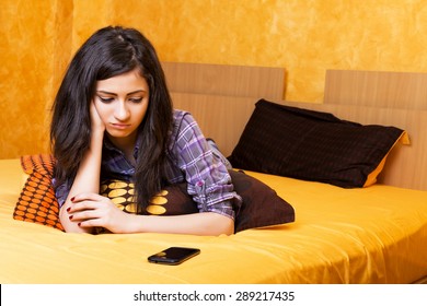 Closeup of pretty teenage girl lying in bed an looking at her mobile phone with sad expression 