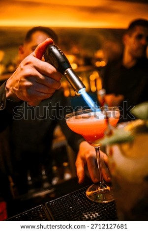 Close-up of a professional bartender using a blowtorch to flame garnish on a red cocktail served in a coupe glass. Dramatic warm bar lighting, shallow depth of field, and dynamic action capture modern mixology, cocktail preparation, and premium nightlife atmosphere. Ideal for concepts of bartending, alcohol beverages, hospitality, luxury bars, and creative drink presentation.