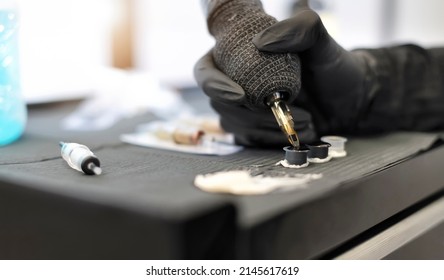Close-up professional female tattoo artist in a black glove with a tattoo machine picks up black ink from a jar - Concept about equipment for making tattoo art - Shutterstock ID 2145617619