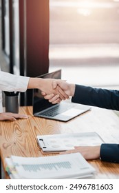 Close-up of a professional handshake between two businesspeople in a modern office setting with documents and a laptop on a wooden desk.