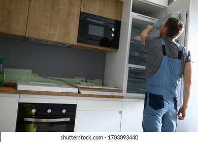 Close-up Of Professional Handyman Repair Fridge. Modern Kitchen Set In New Apartment. Worker In Special Uniform. Renovation Moving Day And Disposable Man Concept