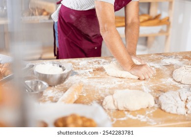 Close-up of professional male baker hands in dark red apron kneading dough on flour-dusted wooden table in artisan bakery, highlighting craftsmanship of traditional breadmaking - Powered by Shutterstock