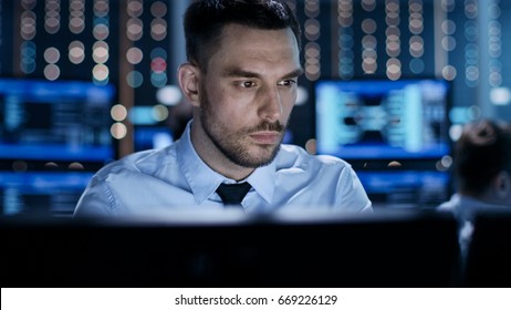 Close-up of a Professional Technical Controller Sitting at His Desk with Multiple Displays Before Him. In the Background His Colleagues Working in System Control Center. - Powered by Shutterstock