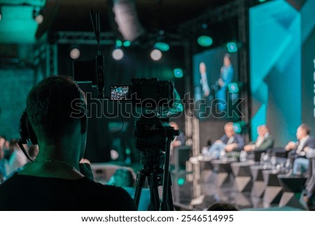 Close-up of professional video camera capturing speakers on stage during business conference. Focus on technology and communication in live event filming.