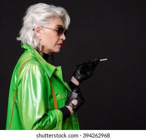 Close-up Profile Of Fashionable Old Woman With Blond Hair Holding Cigarette And Looking Away. Rich Old Woman From Upper Or Elite Class Posing Over Black Background.