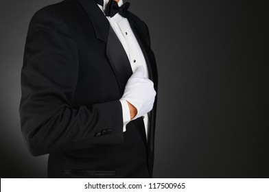 Closeup profile of a man wearing a tuxedo and white gloves holding his lapel. Horizontal format on a light to dark gray background. Man is unrecognizable.