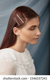 Close-up profile portrait of a dark-haired girl, posing on a blue background. She is wearing white ruffle blouse, pearl earrings and silver chain. Girl has golden flat bobby pin, pearl bobby pin and g