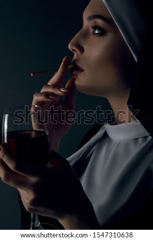 Close-up profile portrait of a nun, posing on a black background. She's wearing dark nun's clothing. The nun is dragging on a cigarette and holding glass of wine in a left hand. 