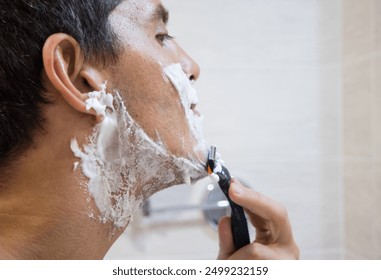 Close-up profile shot of a handsome young white man smiling shaving with his razor in the bathroom at home looking in the mirror removing the shaving foam from his face and starting his skin care rout - Powered by Shutterstock