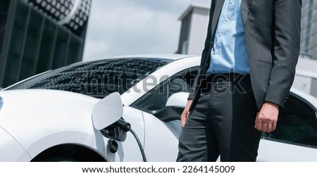 Closeup progressive businessman with electric car recharging at public charging station with background of city residential building. Eco friendly car powered by alternative clean energy.