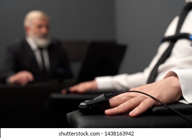 Closeup Of Pulse Indicator On Male Hand During Lie Detector Test. Adult Passing Lie Examination And Answering While Man In Black Smart Suit Controlling Process And Checking Result At Background.