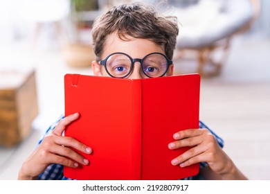 Close-up Of A Quirky Smiling Schoolboy With Glasses, Looking Out From Behind A Notebook And Holding It In His Hands, Sitting At A Table At Home. A Positive Boy Is Happy To Start Classes. Advertising