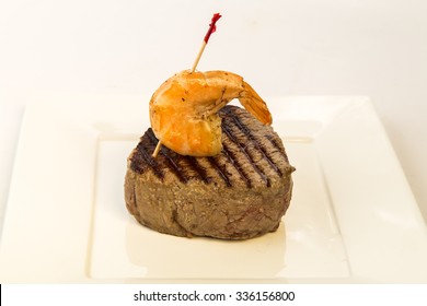 Closeup Of Rare Filet Mignon Topped With Peel And Eat Shrimp On White Square Plate With White Background And Copy Space.