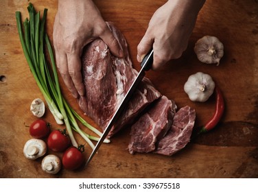 Close-up Of Raw Lamb Meat Lying On Wooden Board With Garlic, Spicy Pepper, Onion, Mushrooms And Tomatoes. Male Chef With Knife Chopping Tenderloin.  