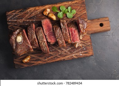 Closeup ready to eat steak new York beef breeds of black Angus with herbs, garlic and butter on a wooden Board. The finished dish for dinner on a dark stone background. Top view