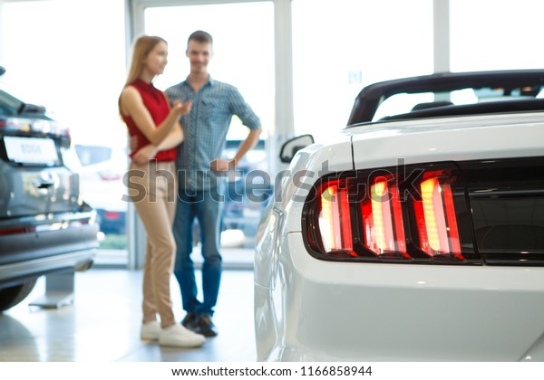 Closeup of rear headlights of white cabriolet in\
auto showroom. Lovely couple discussing and choosing future car on\
background. Young male and female standing near car, gesturing and\
looking at auto.