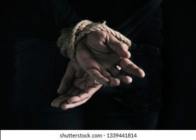 closeup rear view of a man with his hands tied behind his back with rope, against a black background