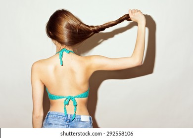 Closeup rear view shot of slim young woman in denim shorts and green crochet bikini with long hair holding and pulling her straight brown hair. Horizontal, retouched, studio shot, intentional shadow.