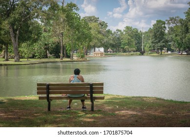 Close-up Rear View A Sportswoman Sitting On A Wooden Bench In Lakeside Neighborhood Park In Houston, Texas, America. Leafy Urban Recreational Space With Mature Trees And A Duck House In Distance