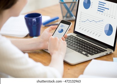 Close-up rear view of young business woman working in office interior on pc holding smartphone and looking at screen with diagrams. Office person using mobile phone and laptop