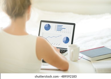 Close-up Rear View Of Young Business Or Student Woman Working At Home Using Laptop Computer, Typing, Looking At Screen With Statistic. View Over The Shoulder. Modern Workplace, Focus On Monitor