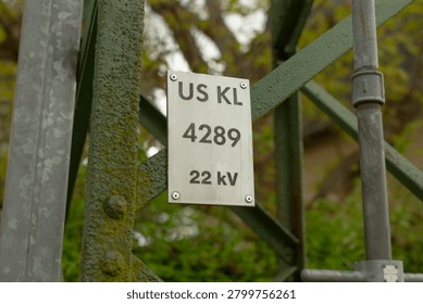 Close-up of a rectangular metal identification plate mounted on a moss-covered green steel lattice support for electrical infrastructure. Plate displays bold