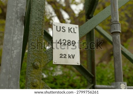 Close-up of a rectangular metal identification plate mounted on a moss-covered green steel lattice support for electrical infrastructure. Plate displays bold
