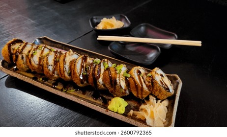 Close-up of a rectangular plate of crispy sushi rolls with a savory sauce, garnished with green onions. Nearby, chopsticks rest next to additional plates of pickled ginger and wasabi.