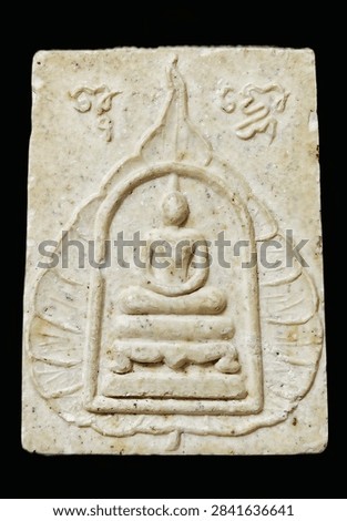 Close-up of a rectangular Thai Buddhist amulet with a raised seated Buddha figure inside an arched shrine, surrounded by leaf-like motifs and engraved script on an aged cream-colored surface.