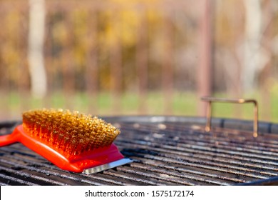 Close-up Of A Red Brush With Golden Bristles And A Scraper For Cleaning A Barbecue Grill Grate. The Concept Of Cleaning After Lunch In Open Air, Picnic, Barbecue, Lunch
