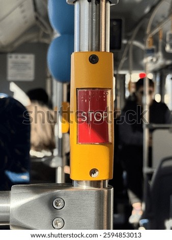 Close-up of a red ‘STOP’ button inside a public bus, with blurred passengers in the background. The image captures the urban transportation experience and daily commute atmosphere.