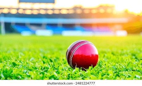 Close-Up of a red cricket ball on a lush green field, with a stadium in the background during sunset. This image captures the essence of cricket and the beauty of the game. - Powered by Shutterstock