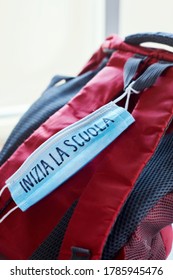 Closeup Of A Red And Gray Bookbag And A Blue Surgical Mask With The Text Back To School Written In Italian, Depicting The Need To Prevent The Infection At School In The Covid-19 Pandemic Situation