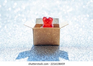 Close-up Of A Red Gummy Bear Candy Peeping Out Of A Small Shipping Box Isolated On A Glittering Background With Copy Space.