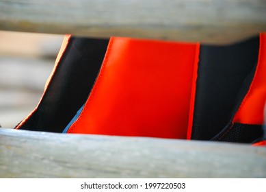 Closeup Of Red Life Jacket With Black Lining Inside Is Hanging Behind White Wooden Balcony. Sea Travel Safety Concept. Empty Space At The Vest Chest Can Use As Mockup.