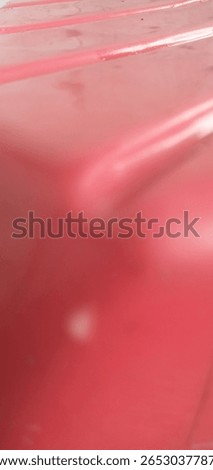 Close-up of a red plastic surface with curved edges and glossy reflections. The image has a soft blur, creating an abstract industrial feel. Subtle lines and light spots add texture.

