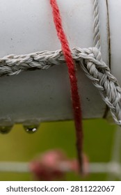 Close-up of red thread woven through gray rope on white surface. Water droplets cling to edges, hinting at rain or dew. Soft focus background shows blurred greenery and pink flower