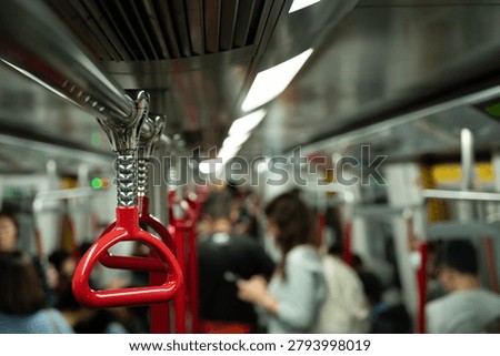 A close-up of a red triangular overhead hanging handle attached to a chrome rail in an MTR train carriage in Hong Kong. The handle is suspended on a coiled chrome fitting. The length of the carriage recedes into the background, with standing and seated commuters, additional red handles, chrome poles, fluorescent ceiling panels, and a reflective metallic ceiling. The carriage is moderately busy with passengers.