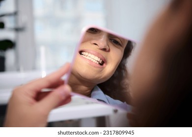 Close-up reflection in the cosmetic mirror of a pretty woman, female patient sitting in dental chair, admiring her beautiful smile and teeth after teeth bleaching procedure in dentistry clinic.