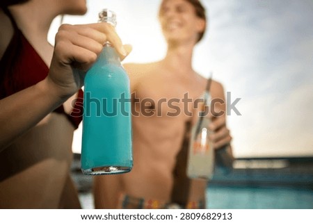 A close-up of a refreshing blue cocktail bottle with a straw in the foreground, with a couple standing out of focus in the background by a resort pool.
