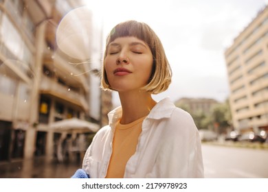 Close-up of relaxed young caucasian woman closing her eyes enjoying walk during day. Brown-haired with bob haircut wears white clothes. Happy weekend concept. - Shutterstock ID 2179978923
