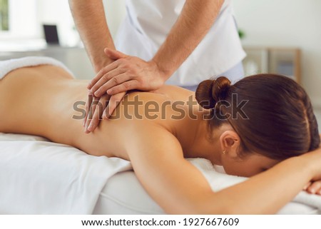 Closeup of relaxed young woman lying face down on massage table and enjoying remedial body massage done by professional masseur in spa salon or wellness center