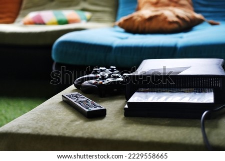 Close-up of remote control, game console and disc packaging on table in living room. Horizontal photo