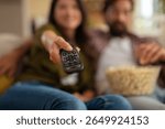 Close-up of a remote control held by a woman with a couple relaxing on a couch, eating popcorn and watching TV at home.